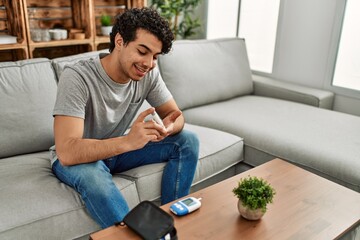 Young hispanic diabetic man measuring glucose sitting on the sofa at home.