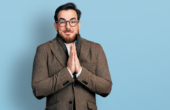 Young hispanic man wearing business jacket and glasses praying with hands together asking for forgiveness smiling confident.