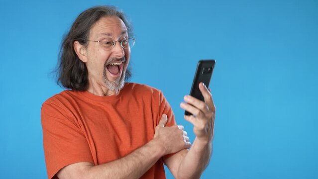 Surprised joyful mature man hold mobile cell phone typing say wow success points to product sale, isolated on solid blue background studio copy space.