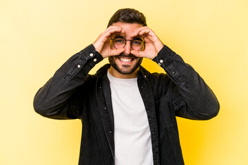 Young caucasian man isolated on yellow background showing okay sign over eyes