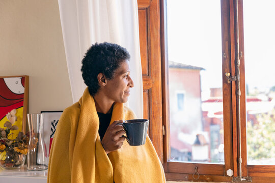 Thoughtful woman with blanket having coffee by window at home
