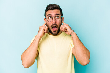 Fototapeta premium Young caucasian man isolated on blue background covering ears with fingers, stressed and desperate by a loudly ambient.