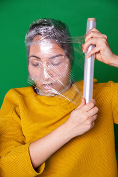 Woman wrapping plastic foil around face against green background