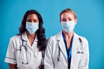 Portrait Of Female Doctor And Nurse Wearing Face Masks Standing In Front Of Blue Studio Background