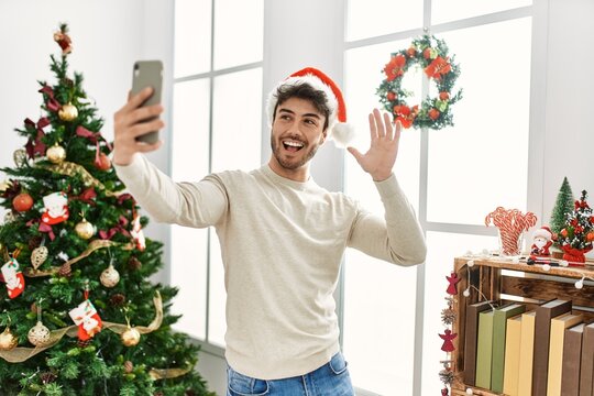 Young hispanic man wearing christmas hat making selfie by the smartphone at home.