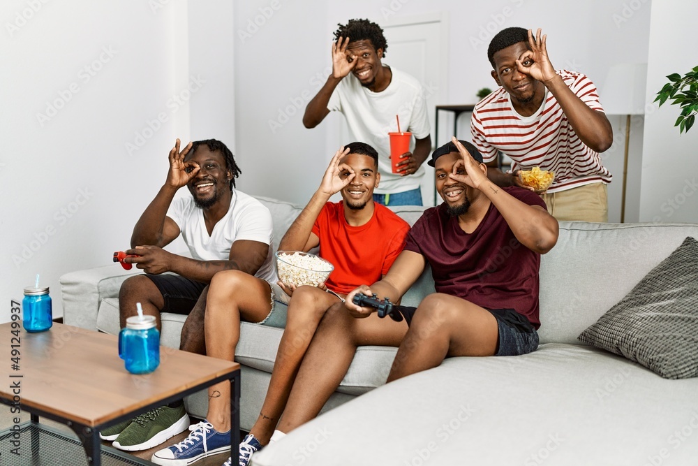 Poster Young african group of friends sitting on the sofa at home playing video games smiling happy doing ok sign with hand on eye looking through fingers