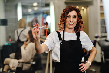 Professional hairdresser looking at the camera while holding a scissor standing in the hair salon.