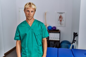 Young blond man wearing physiotherapist uniform standing at clinic depressed and worry for distress, crying angry and afraid. sad expression.