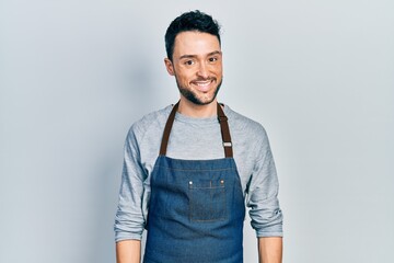 Fototapeta premium Young hispanic man wearing apron and gloves looking positive and happy standing and smiling with a confident smile showing teeth