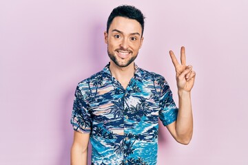 Young hispanic man wearing casual clothes smiling with happy face winking at the camera doing victory sign. number two.