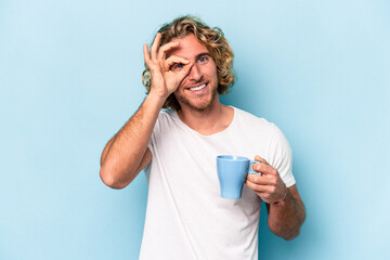 Young man holding a mug isolated on blue background excited keeping ok gesture on eye.