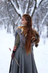 Viking warrior girl with paited face and arrow in her hand dressed in fur coat in winter forest....
