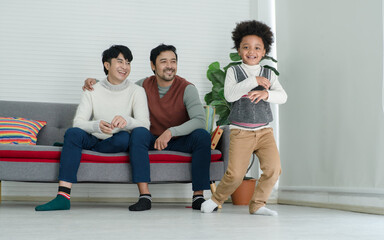 Happy African adopted kid boy wearing sweater dancing Hip Hop in front of Asian young gay parent looking with pride face at living room at home. Lgbt family supporting dream of their son