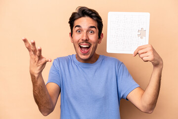 Young caucasian man holding a puzzle isolated on beige background receiving a pleasant surprise,...