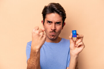 Young caucasian man holding a batterie isolated on beige background showing fist to camera, aggressive facial expression.