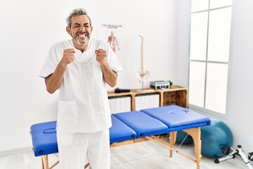 Fototapeta na wymiar Middle age hispanic therapist man working at pain recovery clinic excited for success with arms raised and eyes closed celebrating victory smiling. winner concept.