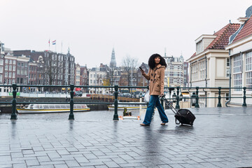 Woman with smart phone pulling wheeled luggage walking on footpath in city