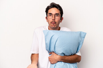Young caucasian man wearing pajamas holding a pillow isolated on white background shrugs shoulders...