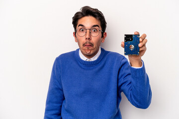 Young caucasian informatic man holding a hard disc drive isolated on white background shrugs shoulders and open eyes confused.