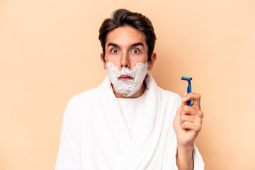 Young caucasian man shaving his beard isolated on beige background shrugs shoulders and open eyes...
