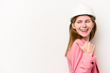 Young architect English woman with helmet isolated on white background points with thumb finger away, laughing and carefree.