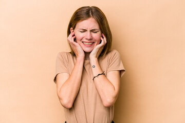 Young English woman isolated on beige background covering ears with hands.