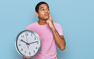 Young handsome hispanic man holding big clock serious face thinking about question with hand on...