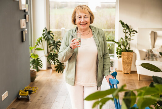 Happy Senior Woman With Glass Of Water Standing By Chair At Home