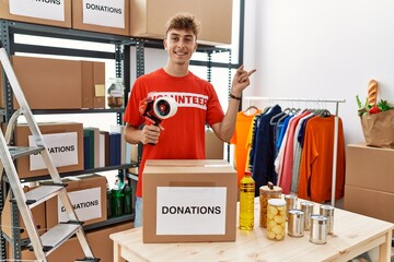 Young caucasian volunteer man packing donations box at charity center smiling happy pointing with hand and finger to the side