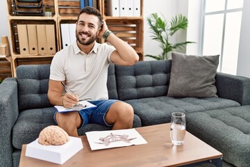 Fototapeta premium Handsome hispanic man working with rorschach test at psychology clinic smiling confident touching hair with hand up gesture, posing attractive and fashionable