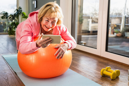 Happy senior woman with smart phone exercising on fitness ball at home