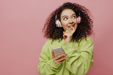 Carefree young caucasian girl puts on headphones, connects smartphone, chooses song indoors. Brunette in green sweater looks up. Technology and lifestyle concept.