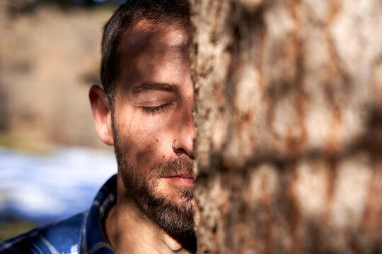 Man with eye closed standing behind tree trunk on sunny day