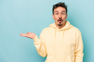 Young caucasian man isolated on blue background impressed holding copy space on palm.