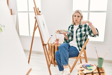Young artist woman painting on canvas at art studio pointing down looking sad and upset, indicating...