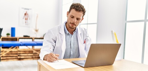 Young hispanic man wearing physiotherapist uniform writing on document at clinic