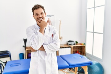 Fototapeta premium Handsome young man working at pain recovery clinic looking confident at the camera smiling with crossed arms and hand raised on chin. thinking positive.