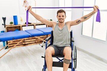 Fototapeta na wymiar Young hispanic man doing rehab using elastic band sitting on wheelchair at clinic
