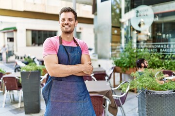 Young hispanic waiter wearing apron standing with arms crossed gesture at coffee shop.