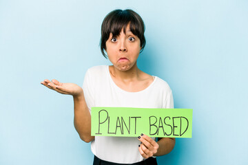 Young hispanic woman holding a plant based placard isolated on blue background shrugs shoulders and...