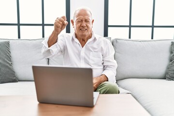 Senior man using laptop at home sitting on the sofa angry and mad raising fist frustrated and furious while shouting with anger. rage and aggressive concept.