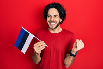 Handsome hispanic man holding estonia flag screaming proud, celebrating victory and success very excited with raised arm