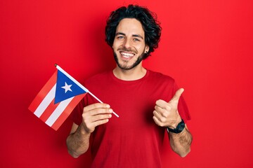 Handsome hispanic man holding puerto rico flag smiling happy and positive, thumb up doing excellent and approval sign