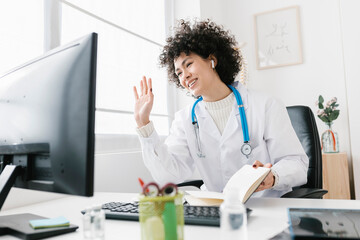 Smiling doctor waving hand on video call through desktop PC in hospital