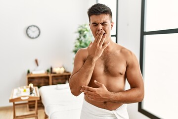Obraz premium Young hispanic man standing shirtless at spa center bored yawning tired covering mouth with hand. restless and sleepiness.