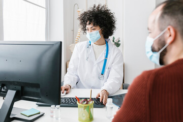 Doctor wearing protective face mask using computer sitting with patient at medical clinic