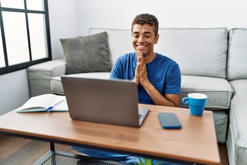 Young handsome hispanic man using laptop sitting on the floor praying with hands together asking for forgiveness smiling confident.