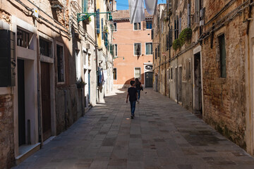 Young tourists walk on a narrow alley of Venice old brick houses on both sides