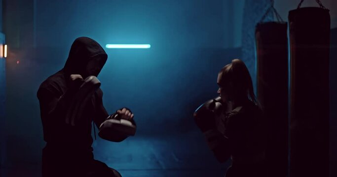 Professional training in fight club, dark room and smoke. Girl fighter practices punches and kicks with a professional trainer. Girl fighter beats boxing paws with her hands in protective gloves. 4k