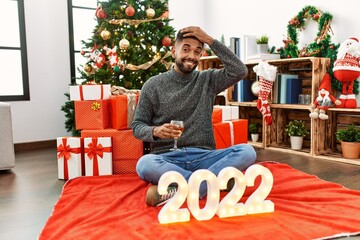 Obraz na płótnie Canvas Young hispanic man with beard sitting by christmas tree celebrating 2022 new year stressed and frustrated with hand on head, surprised and angry face